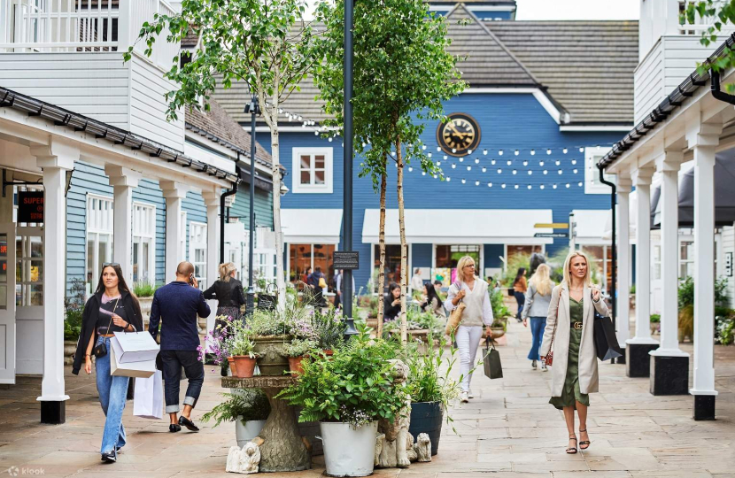 People walking in an outdoor shopping area with shops, plants, and decorations. A blue building is in the background.