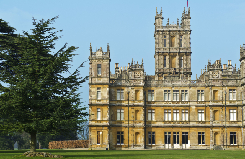 A grand, historic castle with ornate architecture, featuring a central tower and turrets, stands surrounded by a manicured lawn and a large, leafy tree. Bright blue sky in the background enhances the scene.