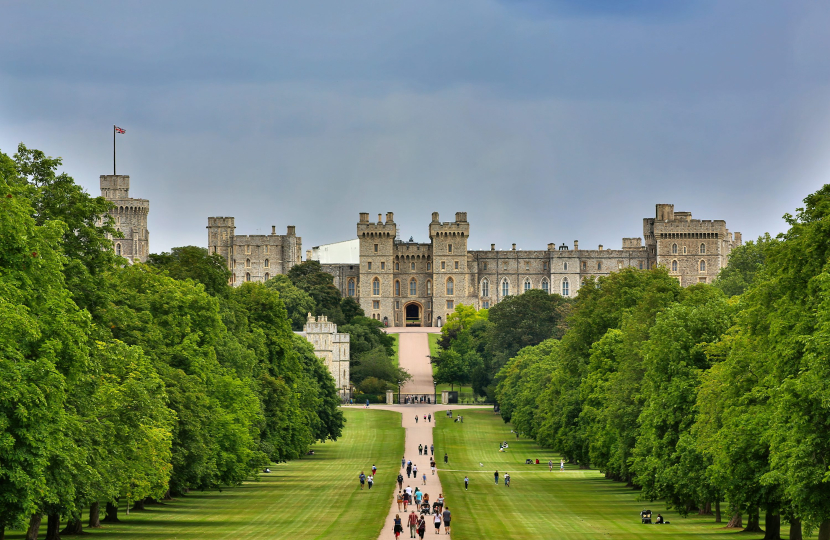 A wide walkway leads to a large castle with a gray stone facade, surrounded by lush green trees and under a cloudy sky. Small groups of people are walking along the path.