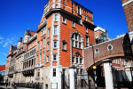 A historic brick building with ornate architectural details and arched windows stands under a clear blue sky. A covered walkway connects two parts of the structure at an upper level. St Mary's Hospital Paddington